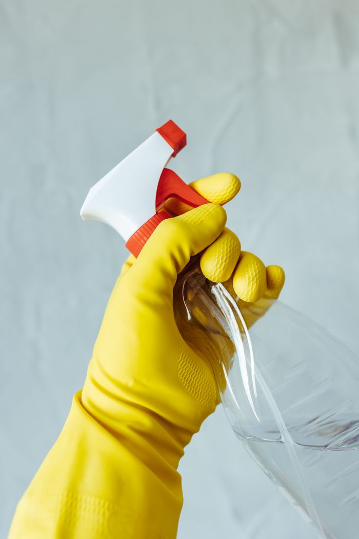 A hand wearing a yellow rubber glove holds a red and white spray bottle against a neutral background.