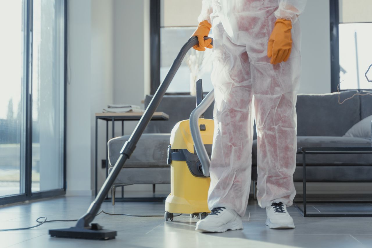 A cleaner in protective coveralls and gloves vacuuming a contemporary living room.